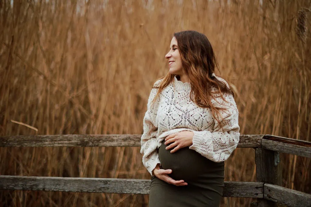 Femme enceinte souriante, pull blanc, devant herbes sèches.