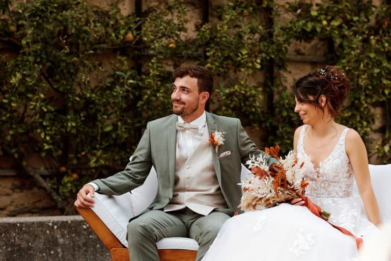 Couple de mariés souriants sur canapé extérieur.