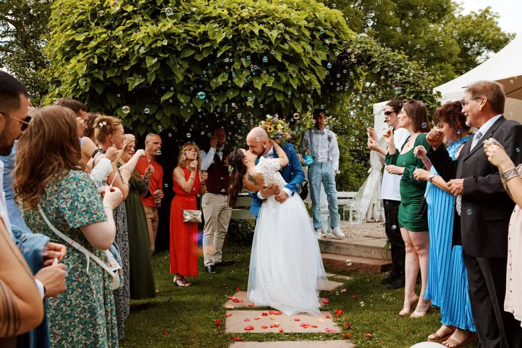 Mariés s’embrassant sous arche fleurie entourés d’invités